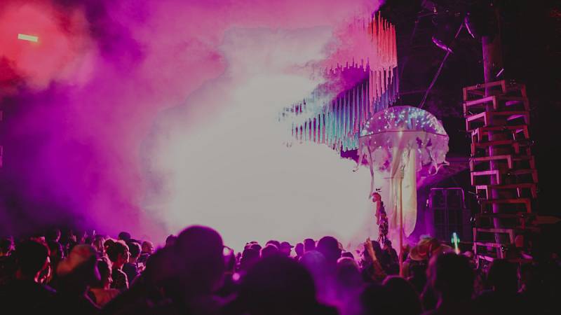 In bunten Nebel gehülltes Publikum vor der Zauberwäldchen-Bühne. Ein Festival-Totem, welches an eine Qualle erinnert, wird langsam vom Nebel verhüllt.