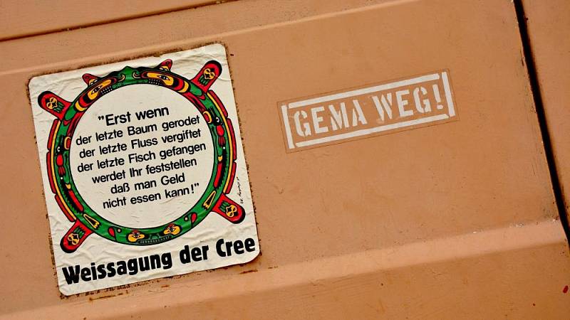 Aufkleber mit der Weissagung der Cree: 'Erst wenn der letzte Baum gerodet, der letzte Fluss vergiftet, der letzte Fisch gefangen ist, werdet ihr feststellen, dass man Geld nicht essen kann.' Daneben ein Graffiti mit der Aufschrift 'GEMA WEG!' auf einer beigen Oberfläche.
